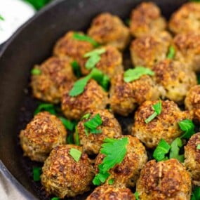 A cast iron skillet filled with browned meatballs garnished with chopped fresh parsley.