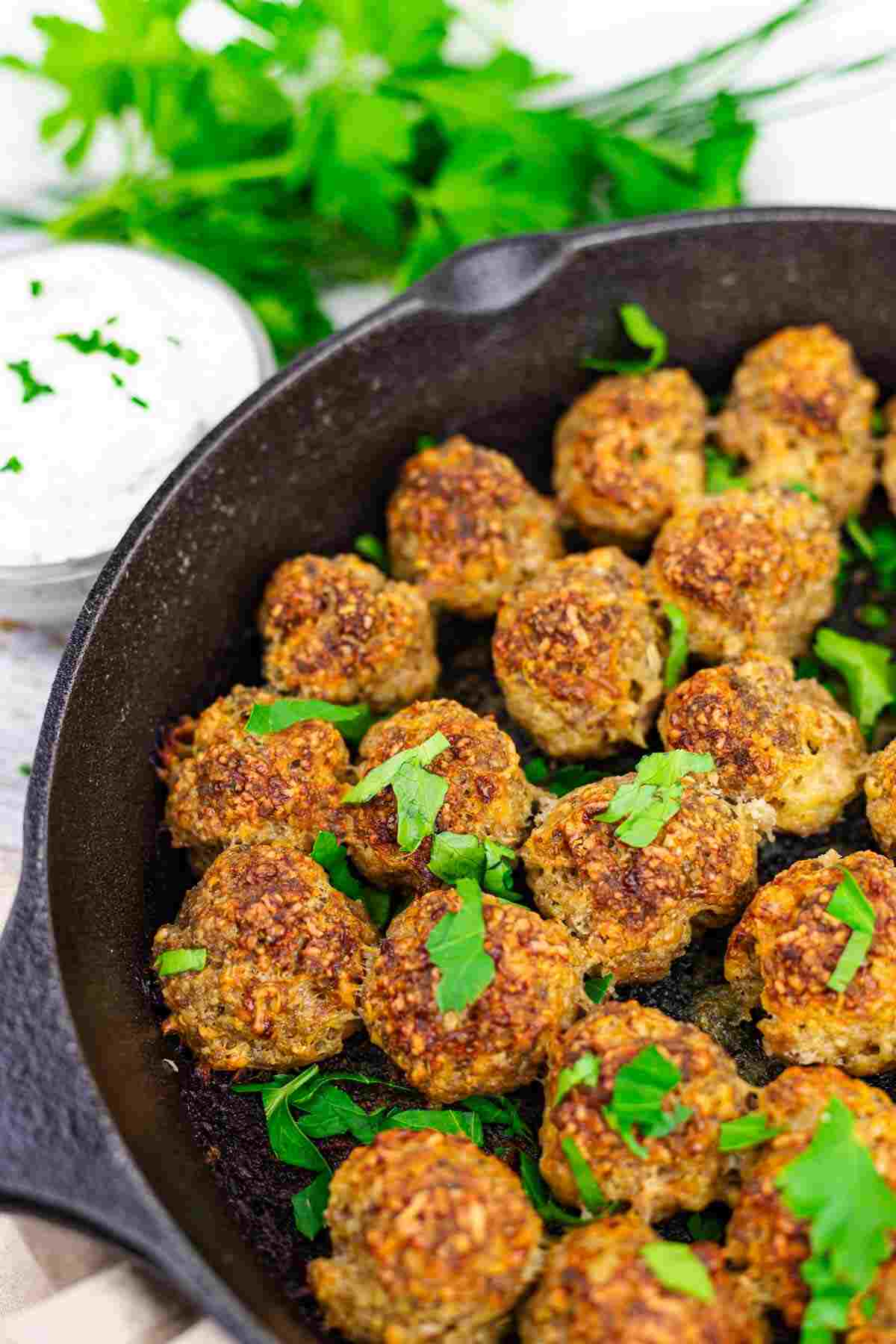 A cast iron skillet filled with browned meatballs, garnished with fresh herbs, with a small bowl of white dipping sauce and parsley in the background.