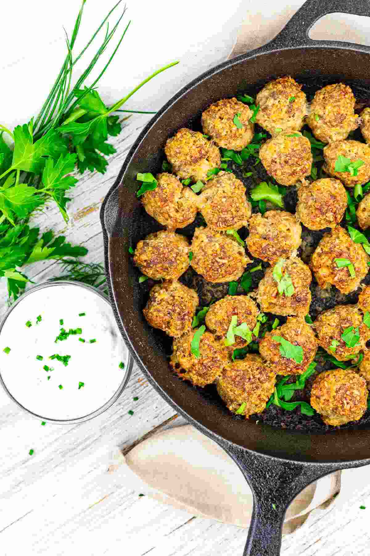 A cast iron skillet filled with cooked meatballs, garnished with chopped herbs, sits next to a bowl of white dipping sauce and fresh parsley on a white wooden surface.