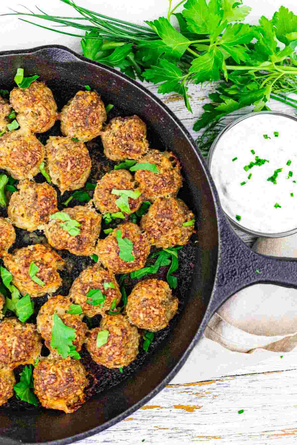 Cooked meatballs garnished with fresh herbs in a cast iron skillet, with a bowl of white dipping sauce and fresh herbs on the side.