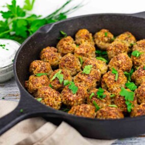 A cast iron skillet filled with cooked meatballs garnished with parsley sits on a napkin, with a bowl of white dipping sauce and fresh herbs in the background.
