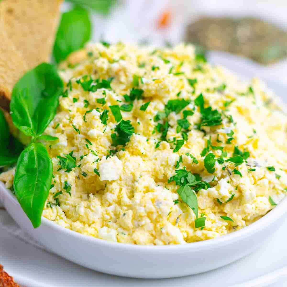 A white bowl filled with crumbled cheese mixed with chopped herbs, garnished with fresh basil leaves, and served with sliced bread.