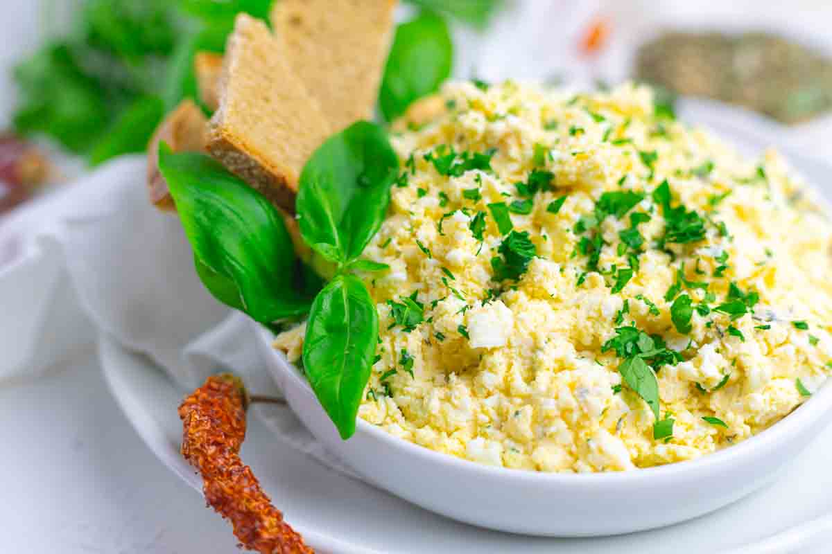 A bowl of baked feta dip garnished with chopped herbs and basil leaves, served with slices of bread on a white plate.