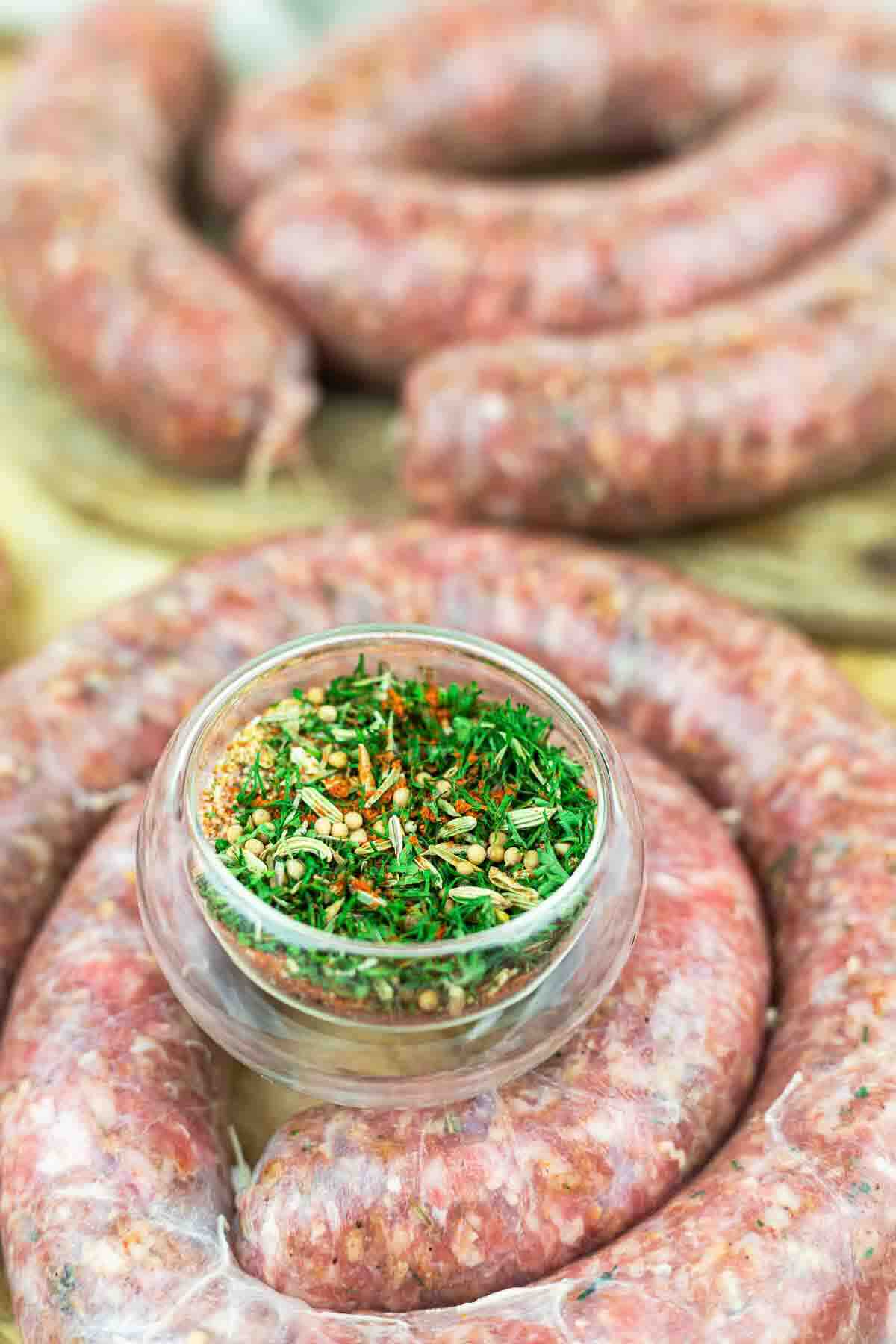 Raw sausage links are coiled on a wooden surface with a small glass bowl of mixed herbs and spices placed on top.