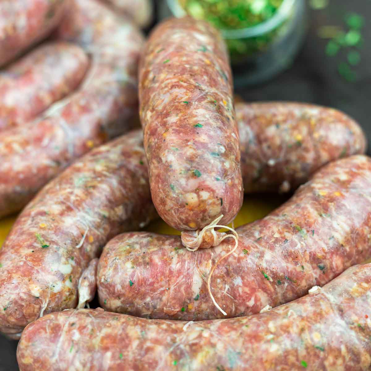 Several raw sausages with visible herbs and seasoning, tied at the ends with string, are displayed on a surface. A blurred jar of herbs is in the background.