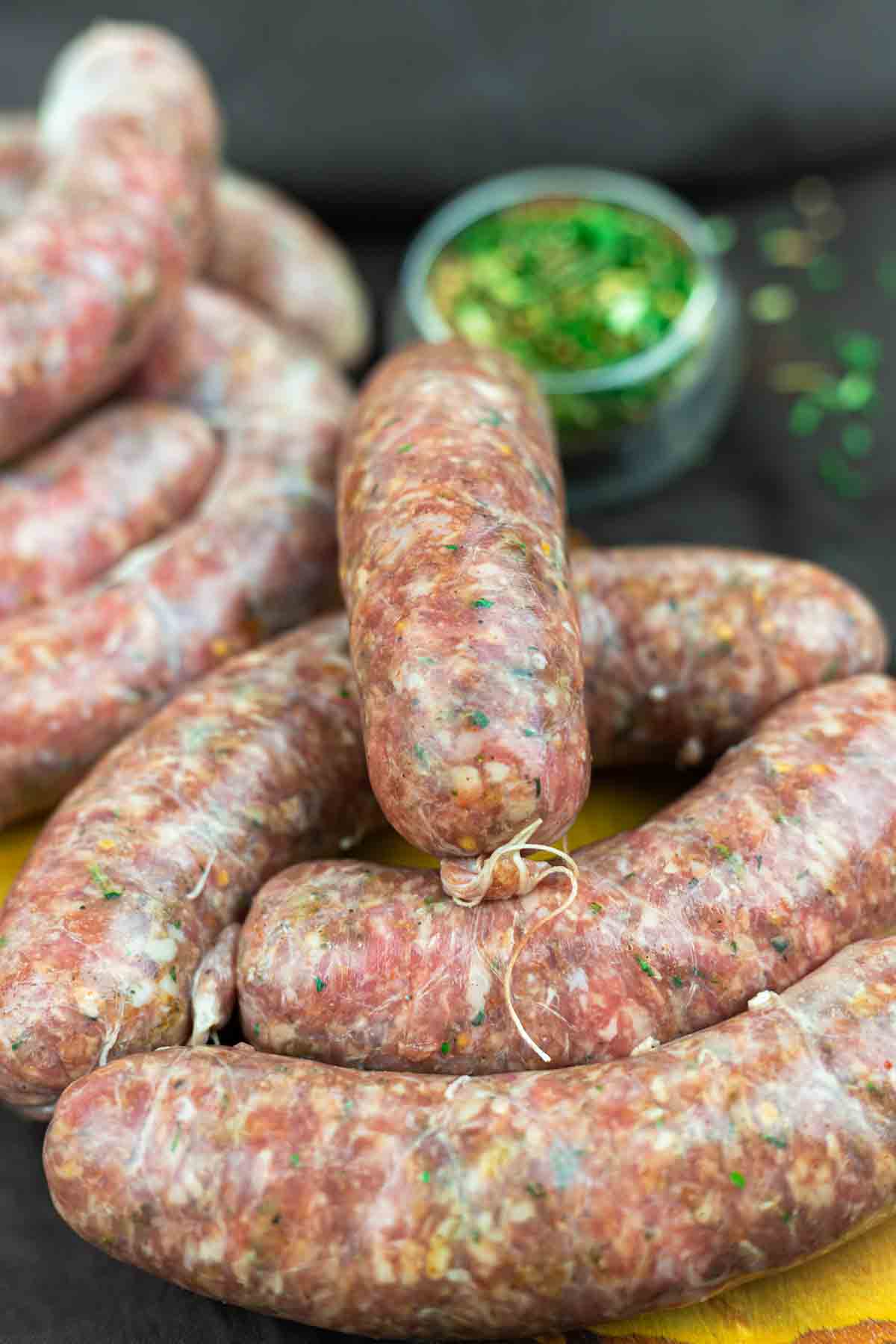 Several uncooked sausages made from ground meat and herbs are arranged on a wooden surface, with a small bowl of chopped green herbs in the background.