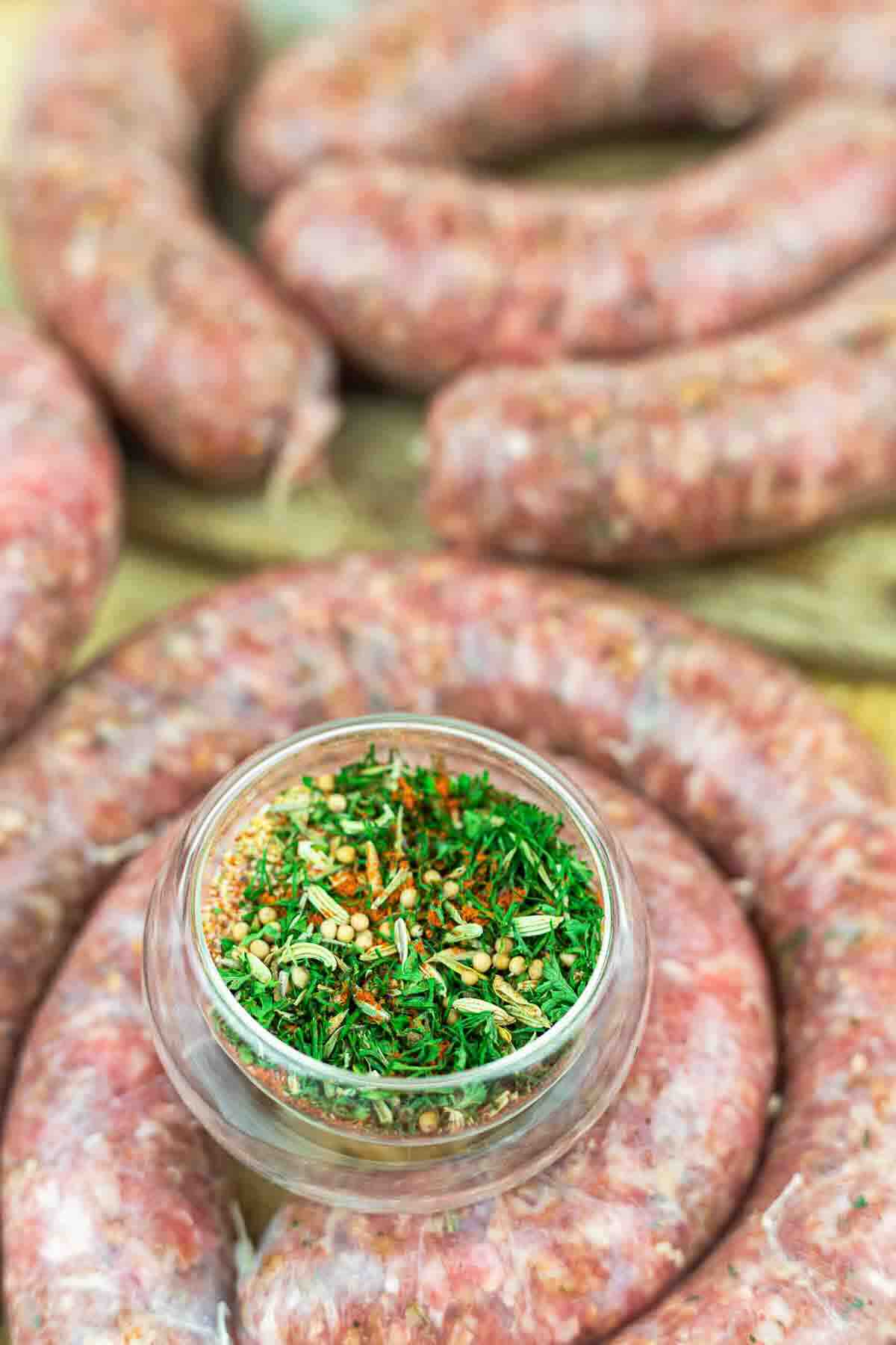A glass bowl of mixed herbs and spices sits on top of coiled raw sausages.
