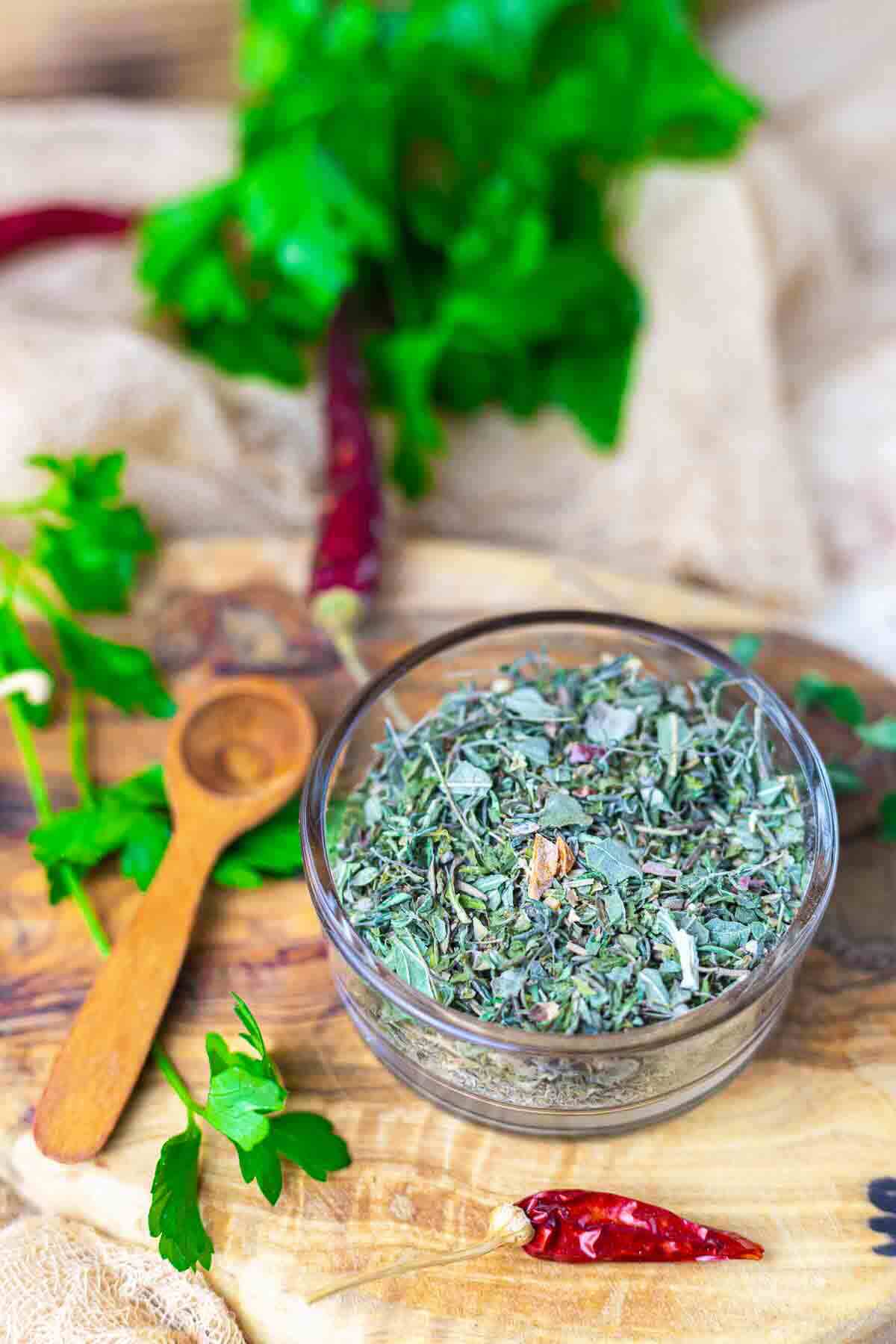 A glass bowl filled with dried mixed herbs sits on a wooden board near a wooden spoon, fresh herbs, and dried red chili peppers.