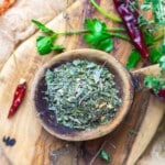 A wooden bowl filled with dried Italian herbs sits on a wooden board, surrounded by fresh parsley and dried red chili peppers.