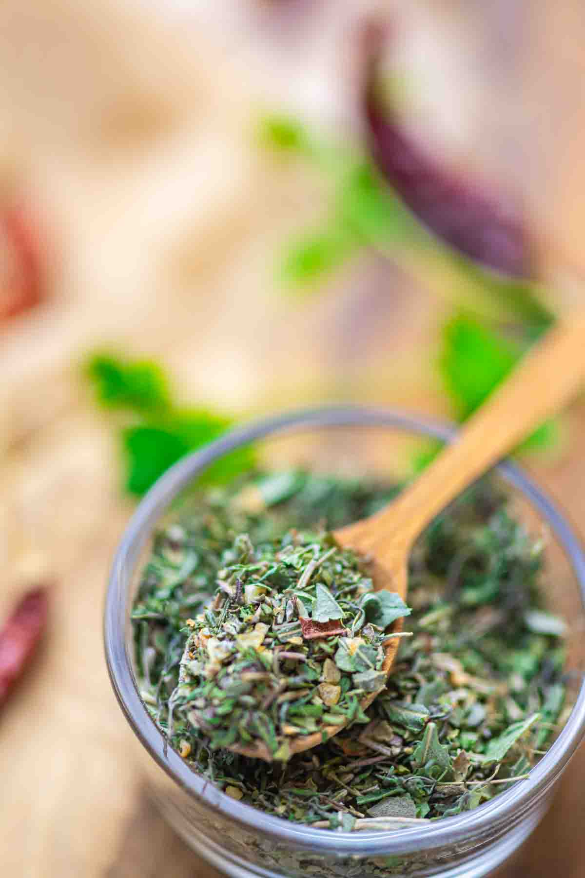 A wooden spoon scoops dried mixed herbs from a small glass bowl, with blurred green and red ingredients in the background.