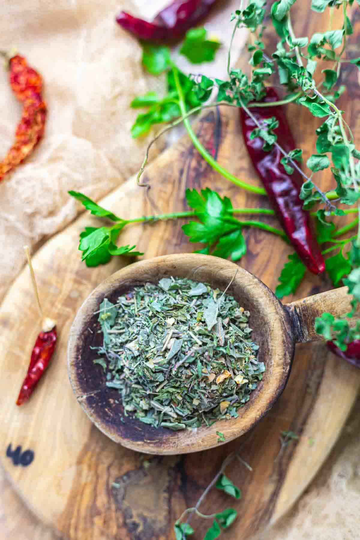 A wooden spoon filled with dried Italian seasoning rests on a wooden board, surrounded by fresh herbs and dried red chili peppers.