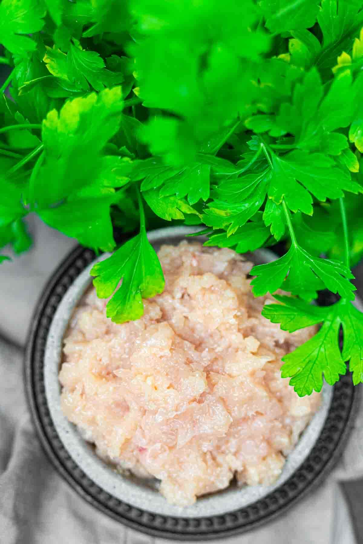 A bowl of raw ground chicken is placed next to fresh parsley leaves.