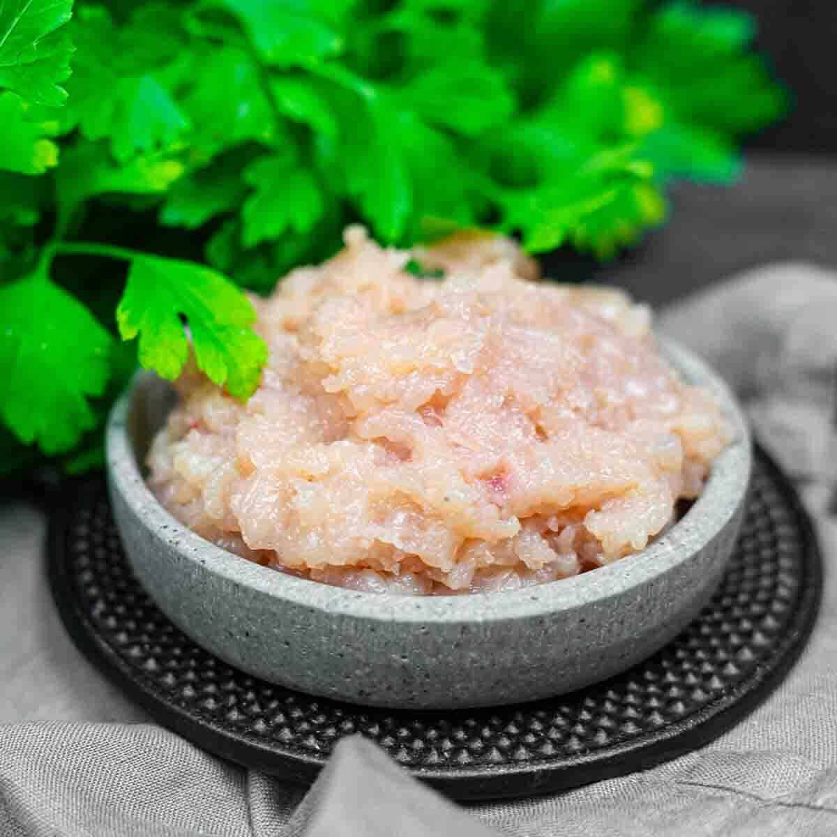 A small gray bowl filled with raw ground chicken sits on a dark mat, with fresh parsley leaves in the background.