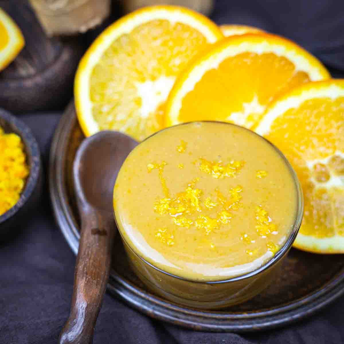 A glass bowl of orange smoothie garnished with orange zest, placed on a dark plate with a wooden spoon and orange slices in the background.