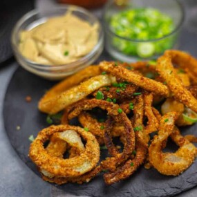 A plate of crispy fried onion rings garnished with chopped chives, served with a bowl of dipping sauce and sliced green onions on the side.