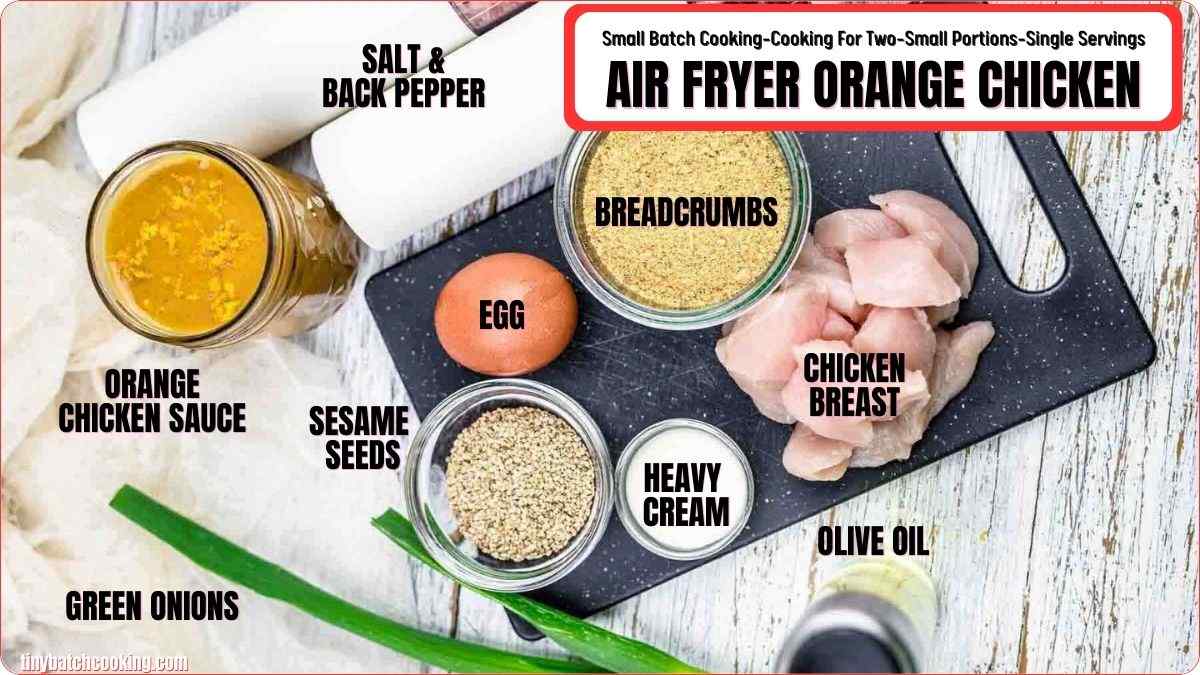 Ingredients for air fryer orange chicken are arranged on a table, including chicken breast, egg, breadcrumbs, orange sauce, green onions, sesame seeds, heavy cream, olive oil, salt, and pepper.
