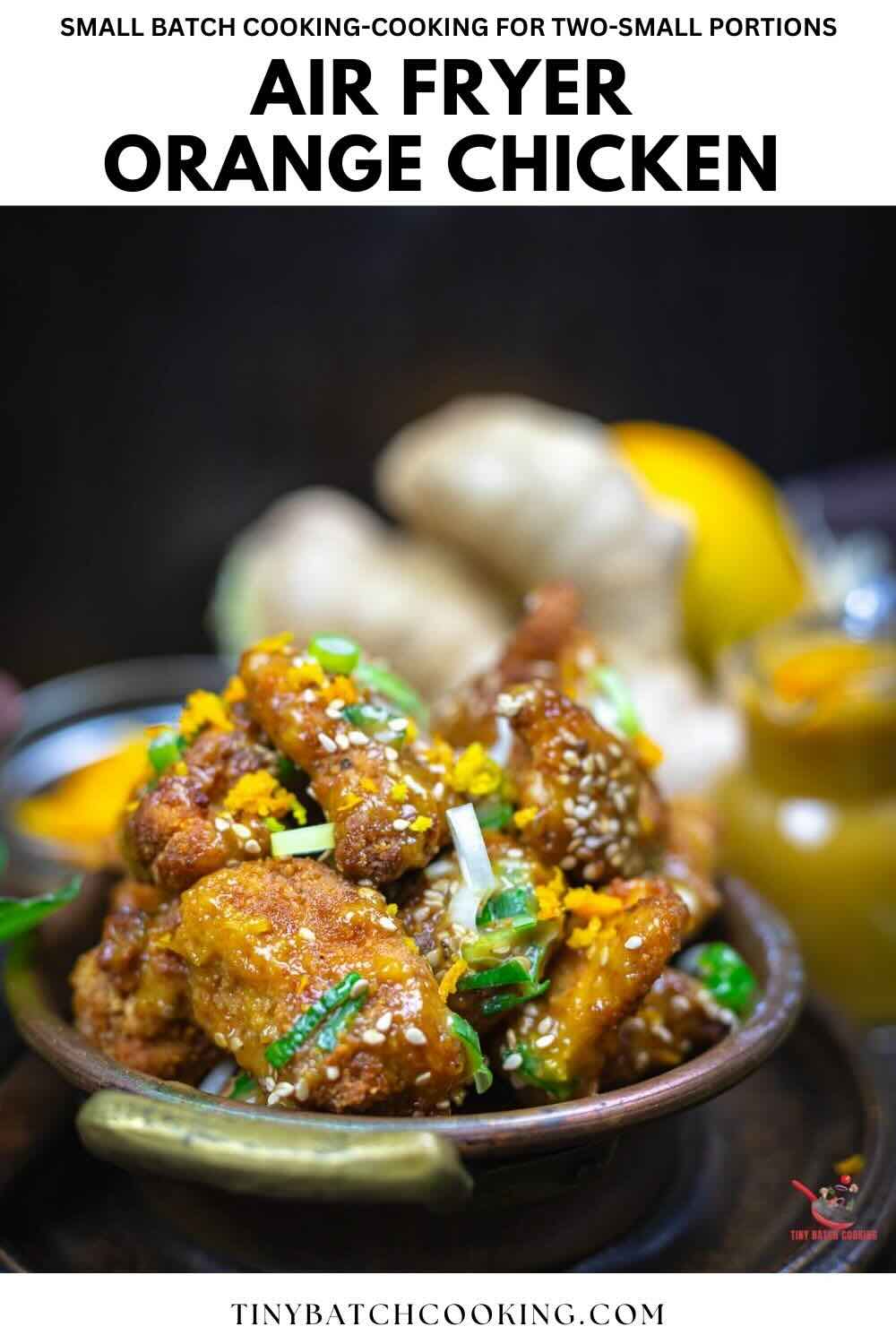 A bowl of air fryer orange chicken garnished with green onions and sesame seeds, with ginger and orange in the background.