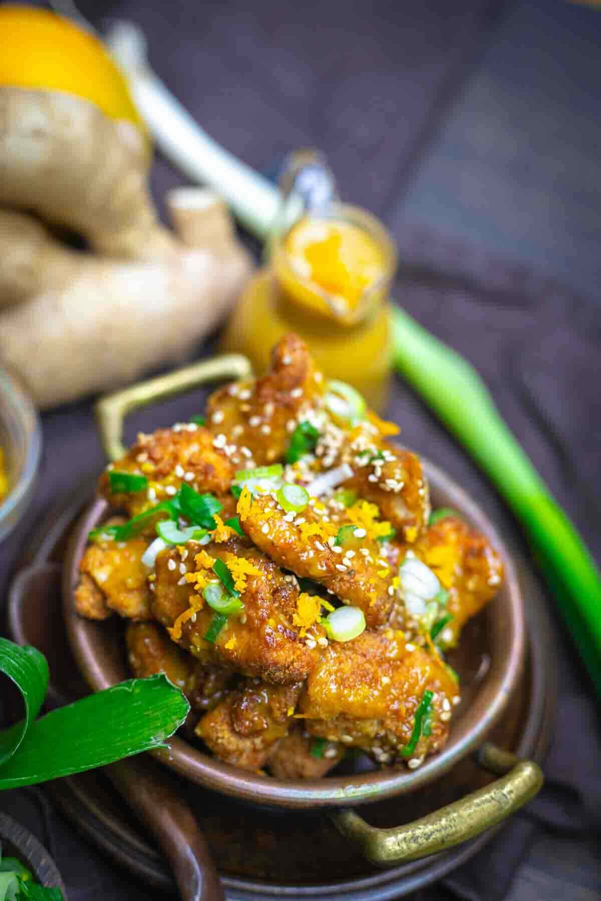 A bowl of glazed chicken wings garnished with sesame seeds and chopped green onions, with ginger root, a green onion, and a small jar of sauce in the background.