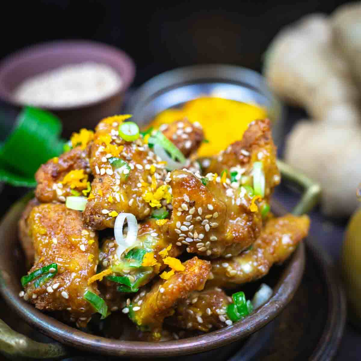 A bowl of crispy fried chicken wings topped with sesame seeds, chopped green onions, and orange zest, with blurred side dishes in the background.