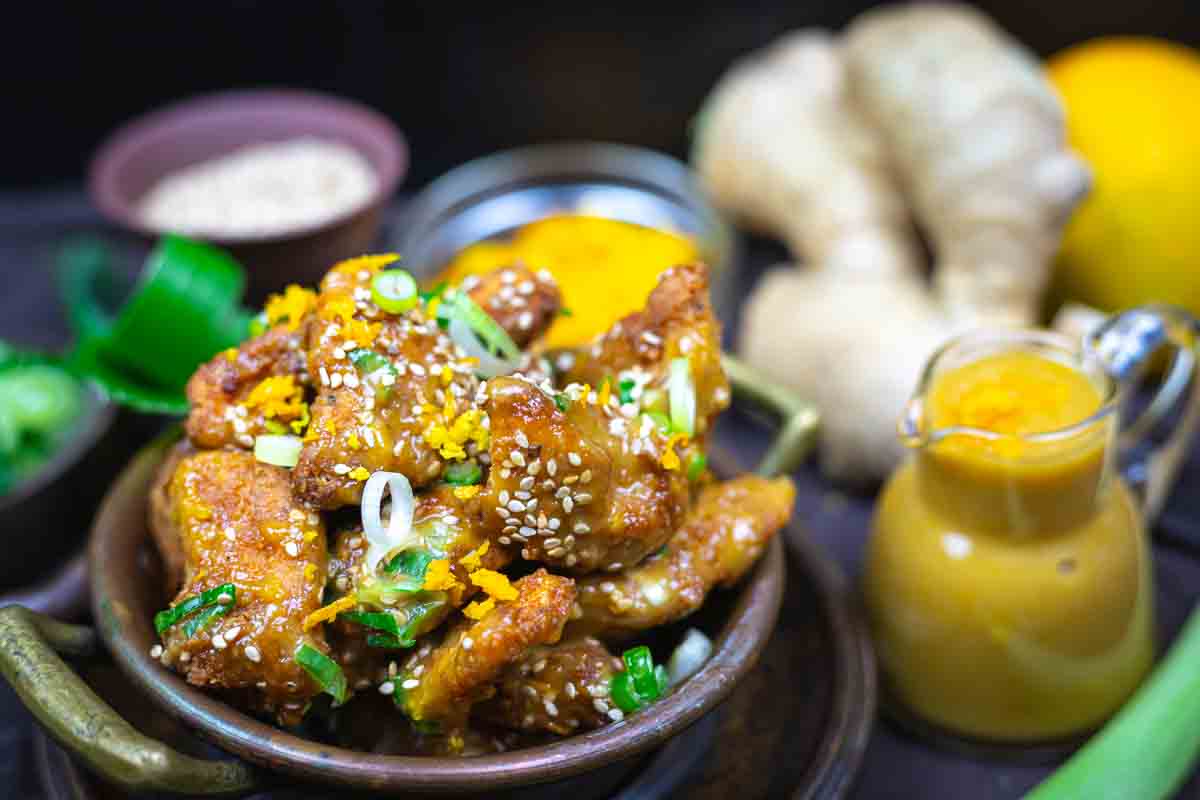 A bowl of orange chicken pieces garnished with sesame seeds and green onions, with sauce and fresh ginger in the background.