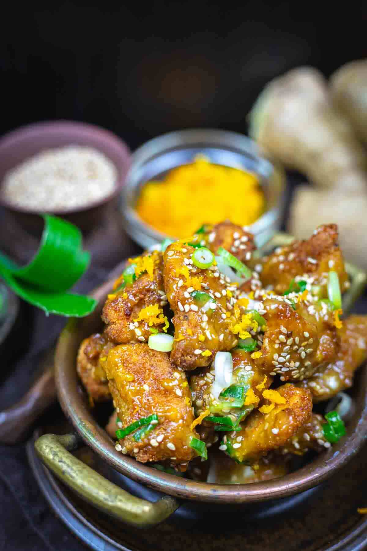 A bowl of crispy fried cauliflower pieces garnished with sesame seeds, chopped green onions, and orange zest, with spices and fresh ginger in the background.