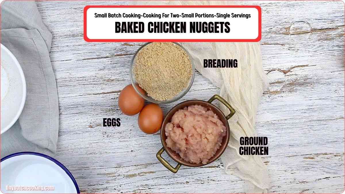 Ingredients for baked chicken nuggets on a white wooden surface: a bowl of breading, two eggs, and a bowl of ground chicken, each labeled.
