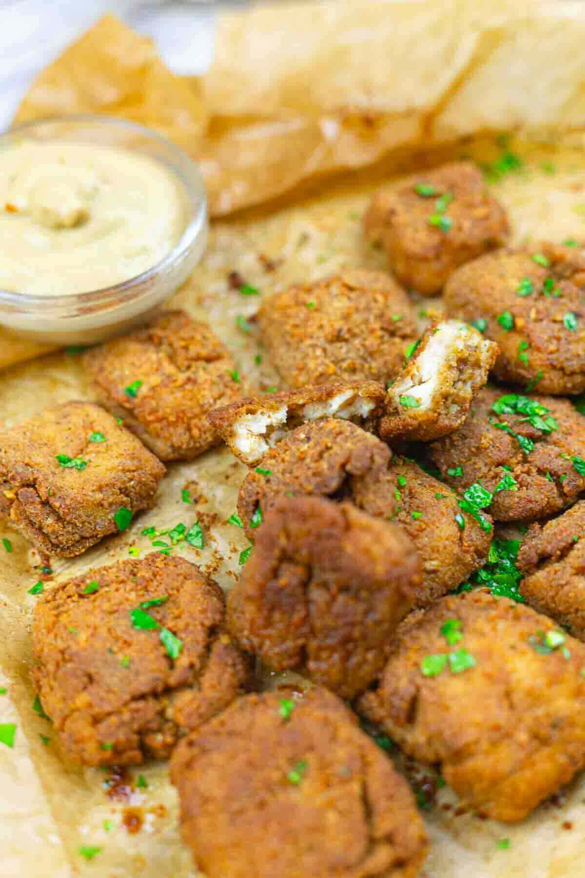 Close-up of several pieces of fried tofu garnished with chopped herbs on parchment paper, with a small bowl of dipping sauce in the background.