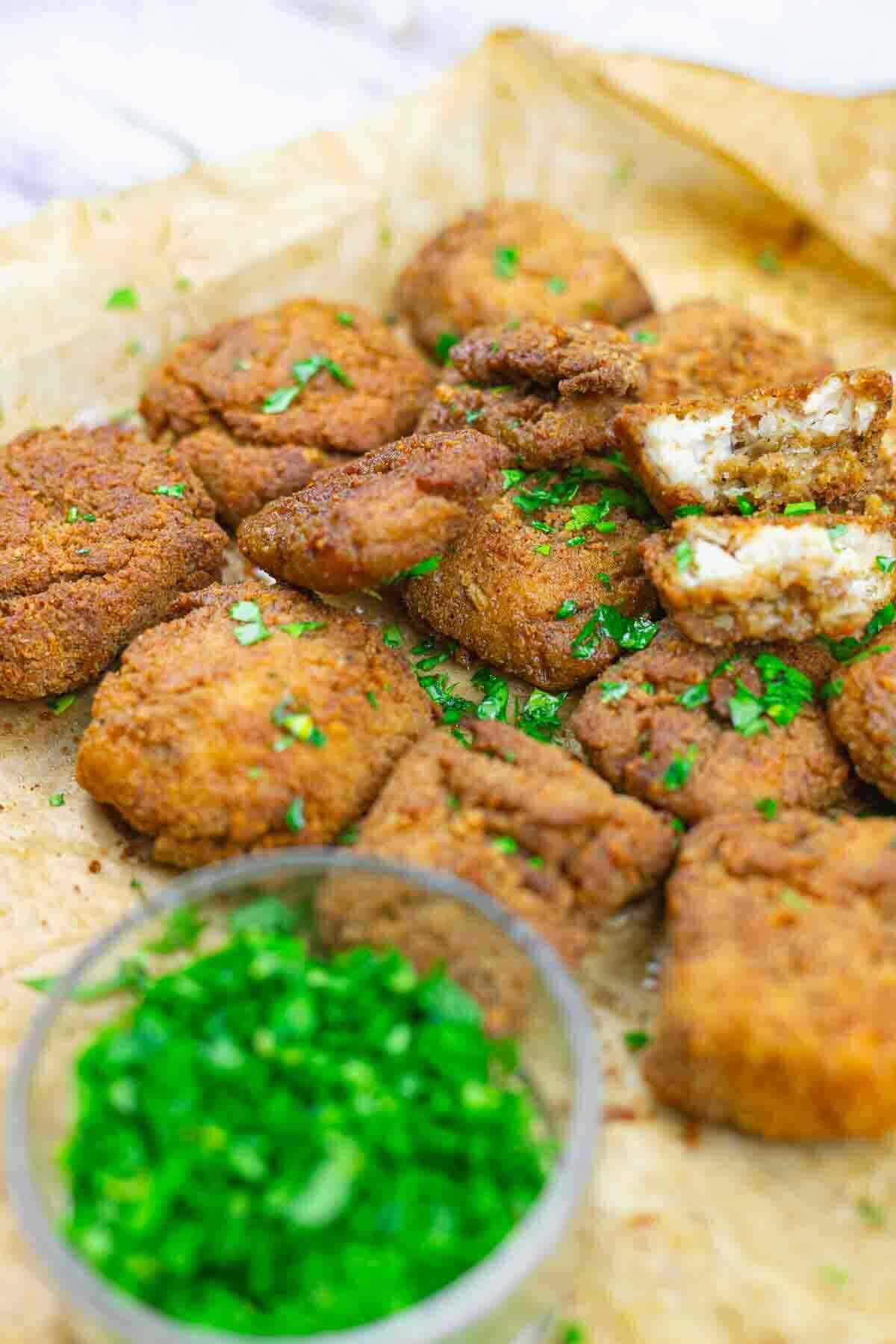 Breaded and fried pieces of food, garnished with chopped parsley, are arranged on parchment paper with a small bowl of fresh herbs nearby.