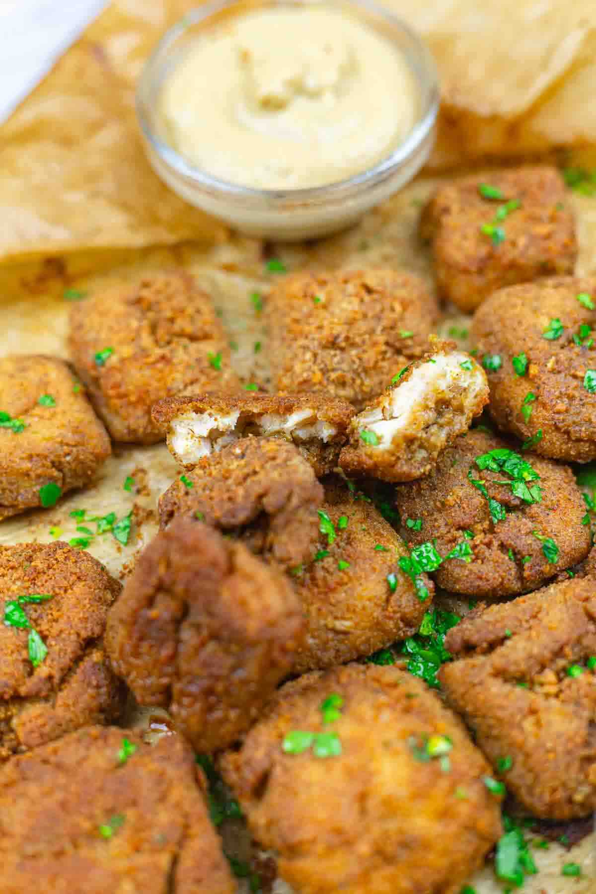 A close-up of fried, breaded tofu cubes garnished with chopped parsley, served on parchment paper with a small bowl of dipping sauce.