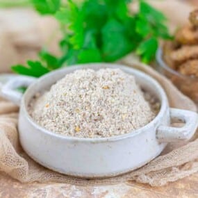 A white ceramic bowl filled with a mound of finely ground seasoning or spice blend, with green herbs visible in the background.