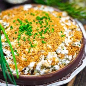 A baked casserole topped with golden breadcrumbs and chopped chives, garnished with fresh chive stalks, served in a round dish on a wooden table.