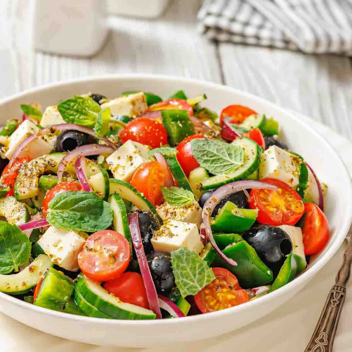 A bowl of Greek salad with tomatoes, cucumbers, red onions, feta cheese, black olives, and fresh mint leaves on a white table.