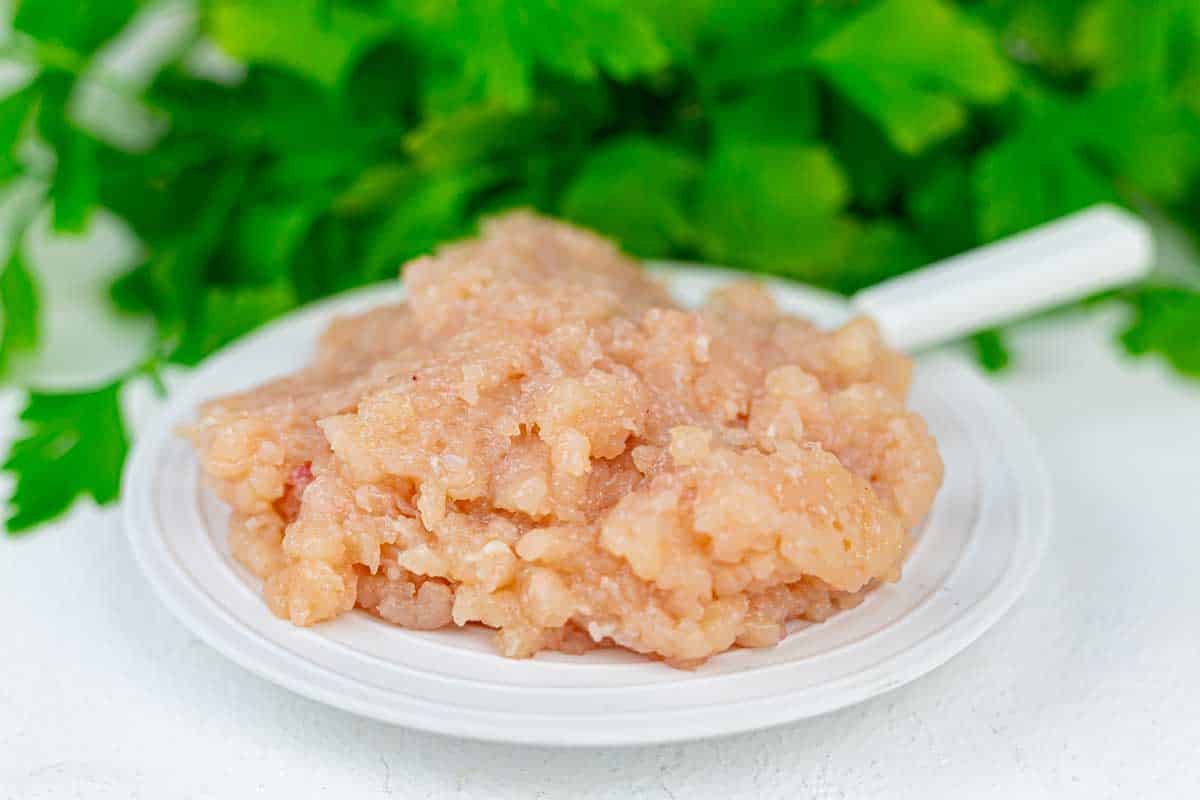 A plate of raw ground chicken with a white spoon, placed on a white surface with green leafy herbs in the background.