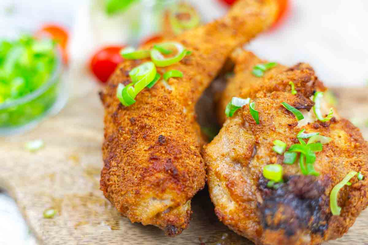 Two pieces of crispy fried chicken garnished with chopped green onions, served on a wooden board with a bowl of sliced green onions in the background.