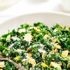 A white bowl filled with a kale salad topped with sliced almonds and grated cheese, with a fork resting on the side.