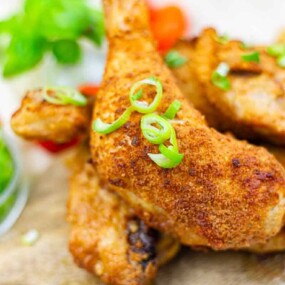 Close-up of crispy fried chicken drumsticks topped with sliced green onions, served on a wooden board with cherry tomatoes and greens in the background.