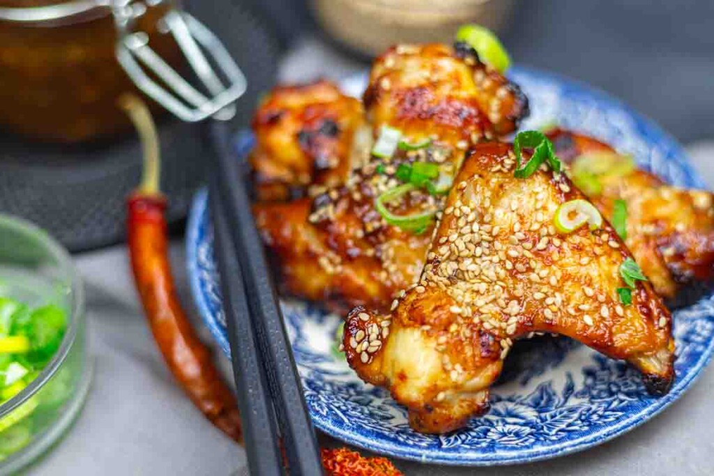 Grilled chicken wings topped with sesame seeds and chopped green onions, served on a blue patterned plate with chopsticks.