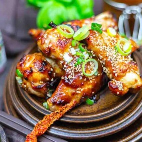 A plate of glazed chicken drumsticks garnished with sliced green onions and sesame seeds, surrounded by chopsticks and tableware.