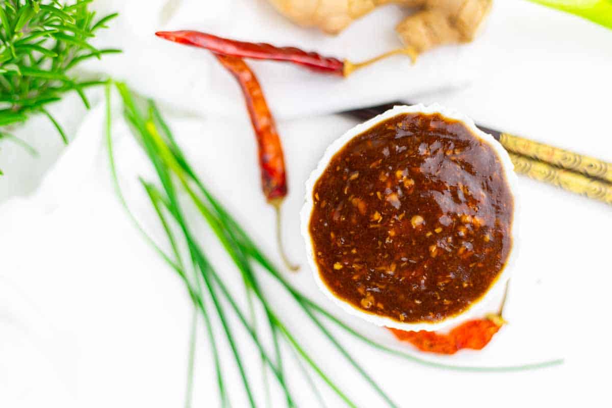 A bowl of spicy BBQ sauce sits on a white surface, surrounded by dried red chili peppers, ginger, herbs, and green chives.