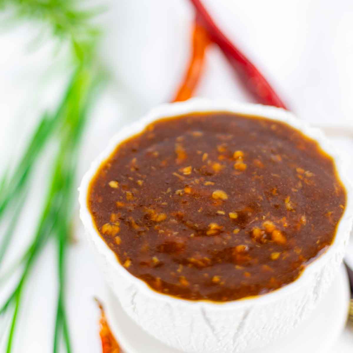 A white bowl filled with a thick, chunky brown sauce, with herbs and dried red chilies in the blurred background.