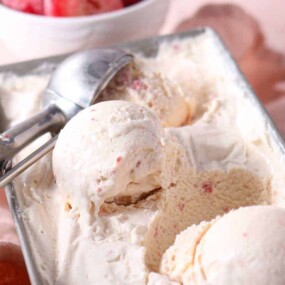 A metal container of vanilla ice cream with an ice cream scoop and three scoops, set on a light pink cloth with a bowl of strawberries in the background.