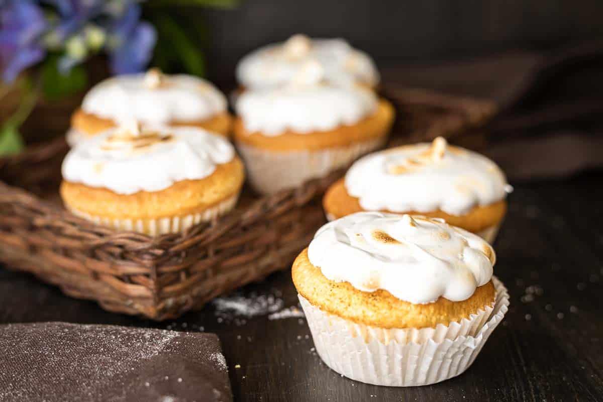 Five cupcakes with white frosting are arranged on a dark surface and in a wicker tray, with a brown napkin and scattered flour nearby—perfect easy desserts as summer ends.
