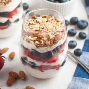 A glass filled with layers of yogurt, strawberries, blueberries, and granola sits on a white surface next to a spoon, whole strawberries, almonds, and scattered blueberries.