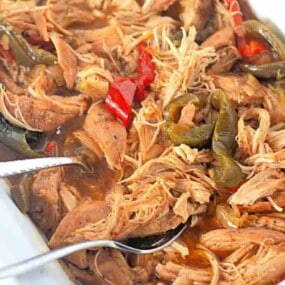 Shredded seasoned meat with sliced red and green peppers in a white dish, with metal serving utensils.