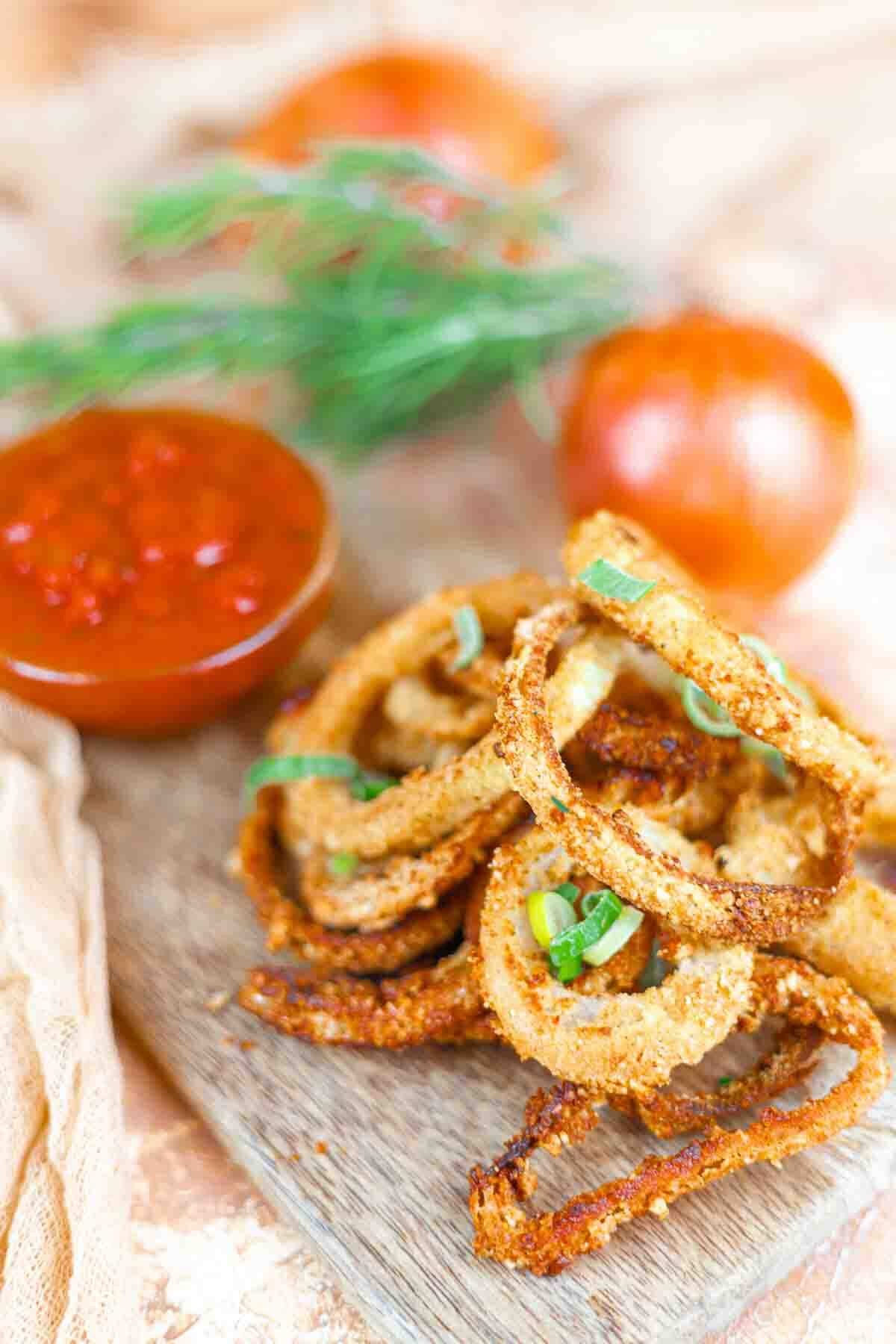 A pile of crispy onion rings garnished with green herbs on a wooden board, served with a bowl of red dipping sauce.
