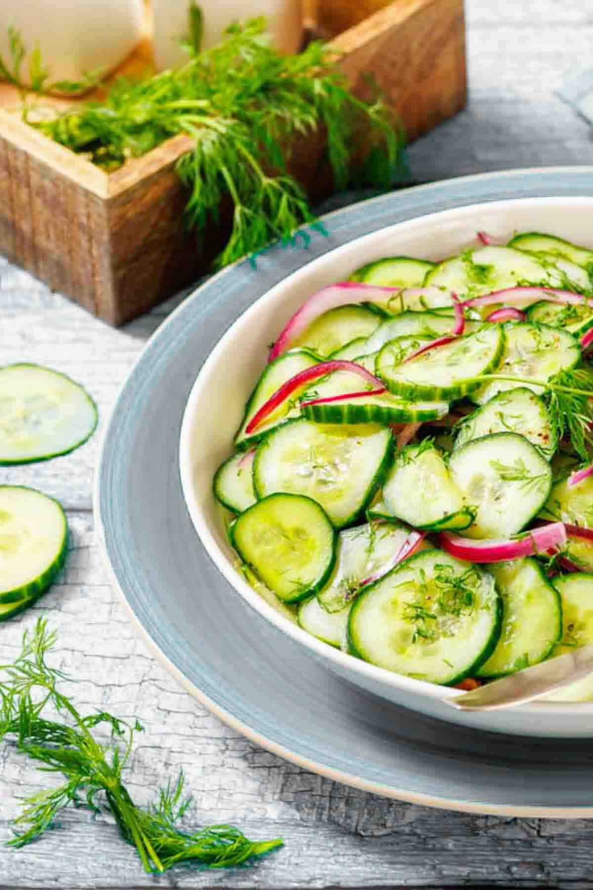A bowl of cucumber salad with sliced cucumbers, red onions, and dill, placed on a gray plate with fresh dill and a wooden box in the background.