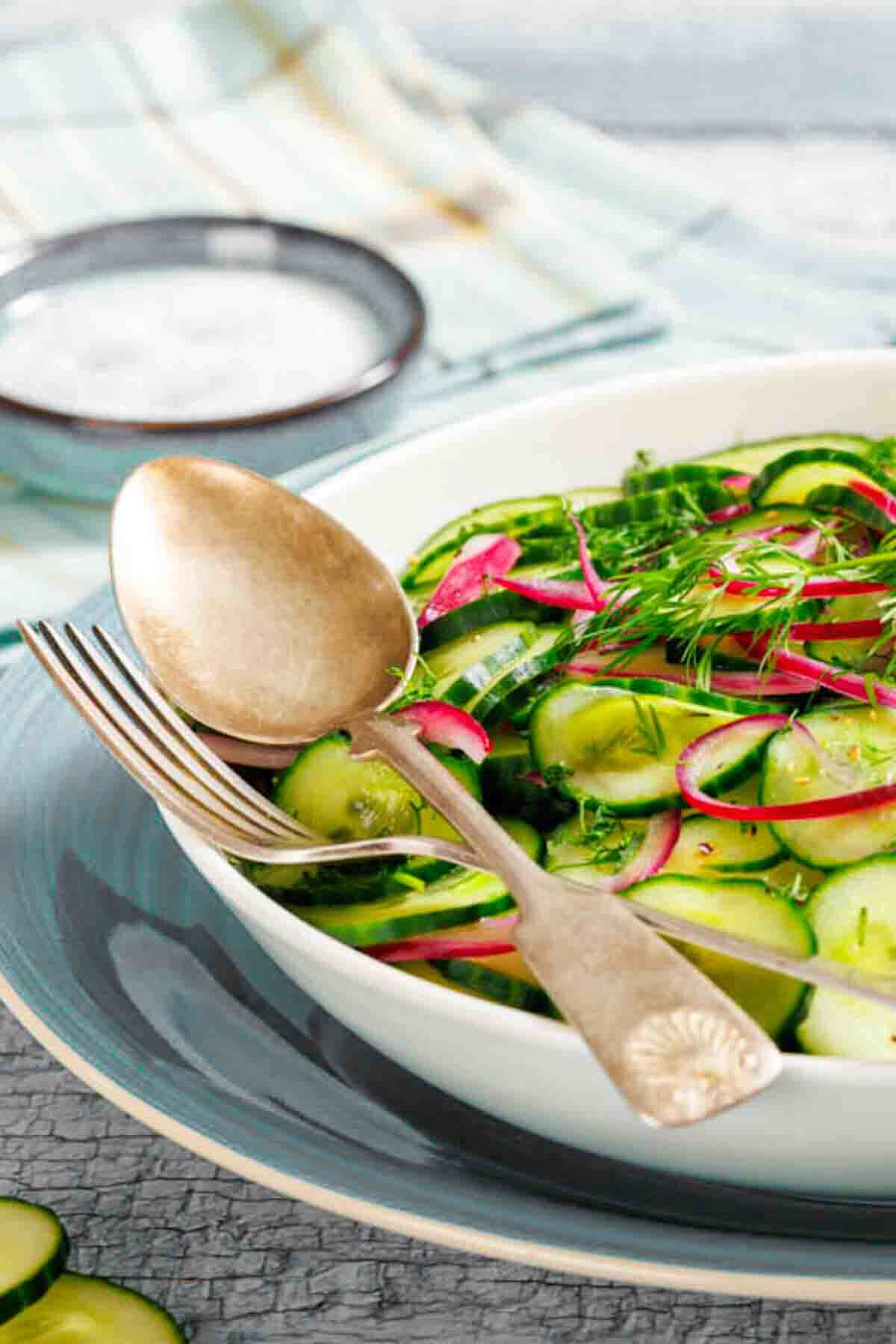 A bowl of cucumber and radish salad garnished with fresh herbs, with a fork and spoon resting on the edge of the bowl.
