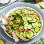 A bowl of cucumber and red onion salad garnished with fresh dill, with a fork and spoon resting on the side of the bowl.
