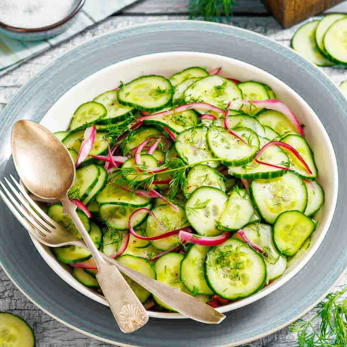 A bowl of cucumber and red onion salad garnished with fresh dill, with a fork and spoon resting on the side of the bowl.