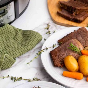 A plate with slices of pot roast, baby potatoes, and carrots, garnished with herbs. A slow cooker, green towel, and sliced roast on a cutting board are nearby.